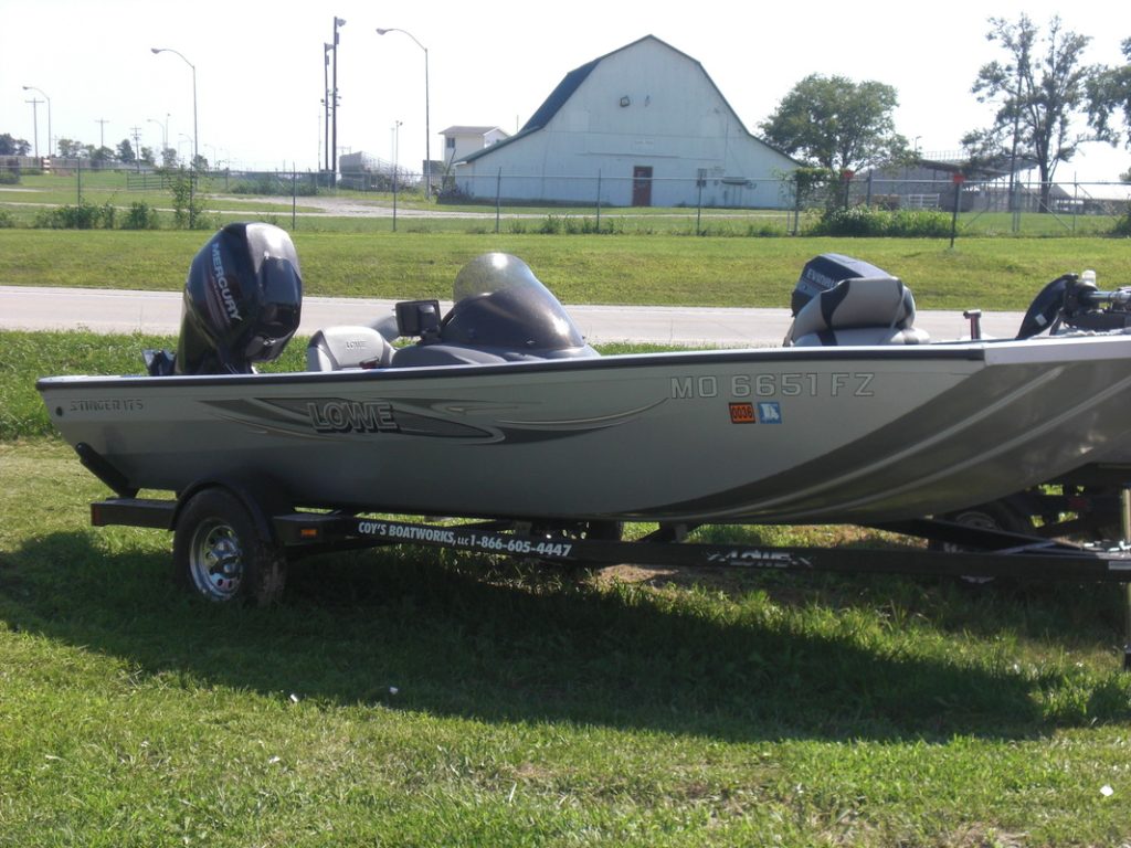A silver Lowe fishing boat on a trailer is parked on grass beside a road, with a large white building in the background.