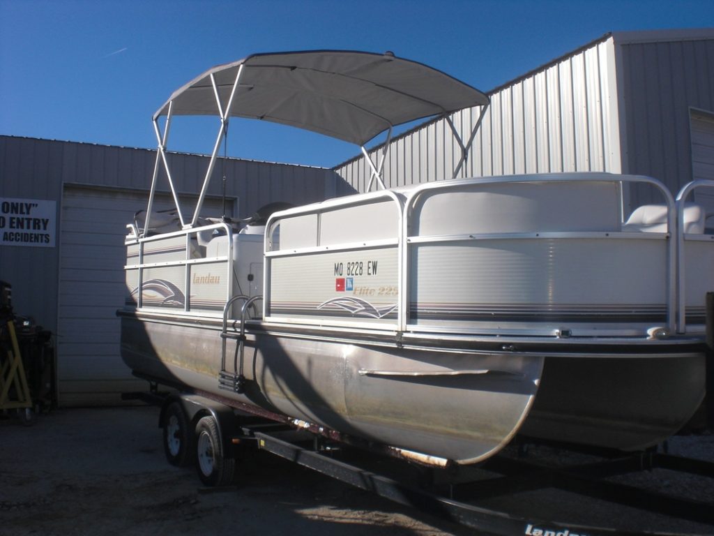 White pontoon boat with a canopy is parked on a trailer in front of a metal building under a clear blue sky.