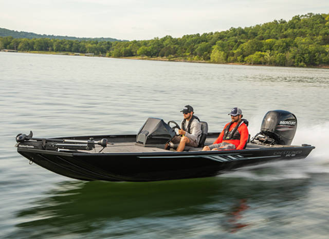 Two men ride a black motorized fishing boat on a lake, with trees and hills visible in the background.